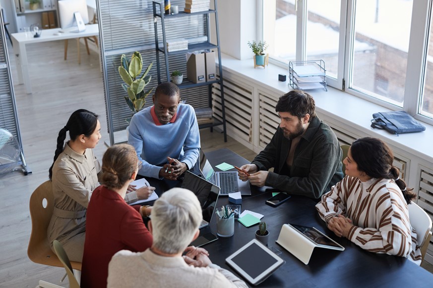 Six diverse group members meeting and discussing in an office while using laptops and tablets